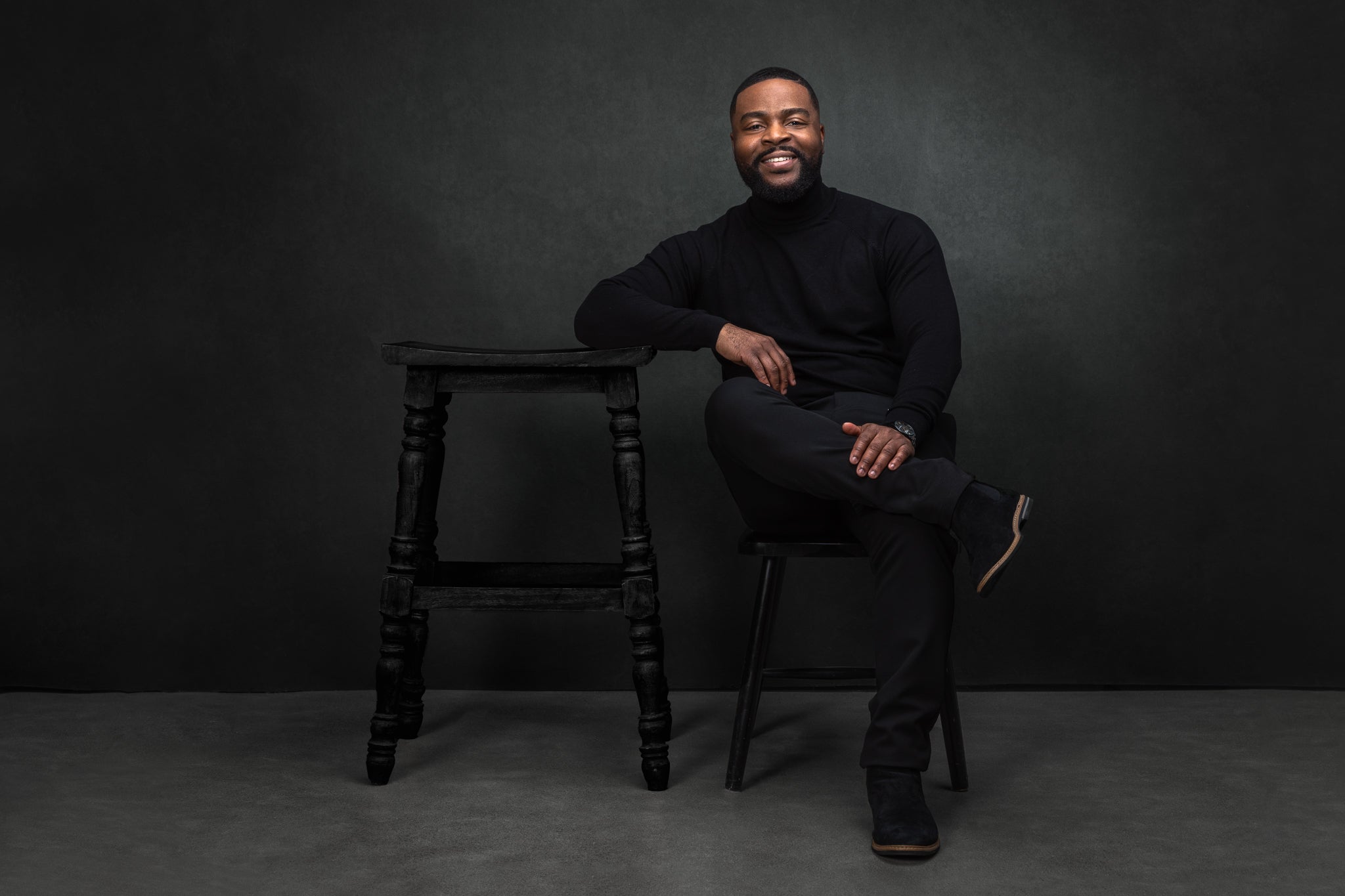 Smiling man dressed in black sitting on a chair with a wooden stool beside him against a dark background