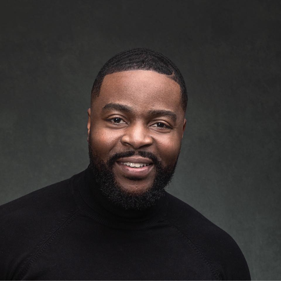 Smiling man with short hair and beard wearing black turtleneck sweater against dark background